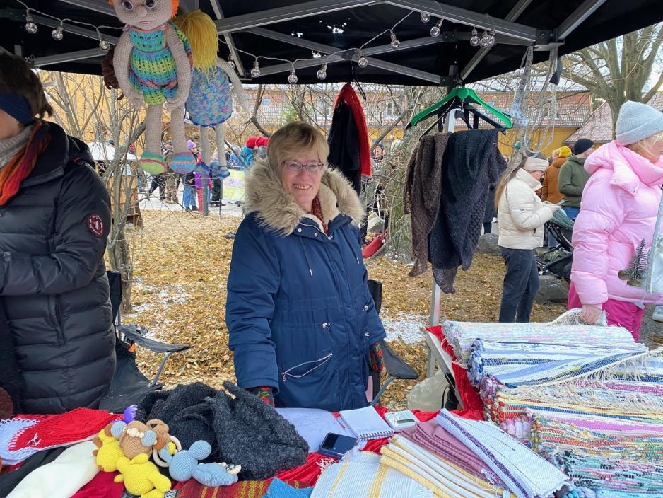 Bildet viser eldre dame som smiler bredt til kameraet. P&aring; boden foran henne ligger det mye forskjellig strikket&oslash;y, dukker, og filleryer.
