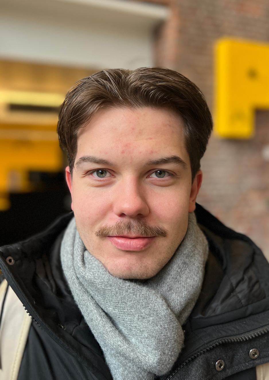 Mann med brunt h&aring;r i midtskille, bart og n&oslash;ytralt ansiktsuttrykk