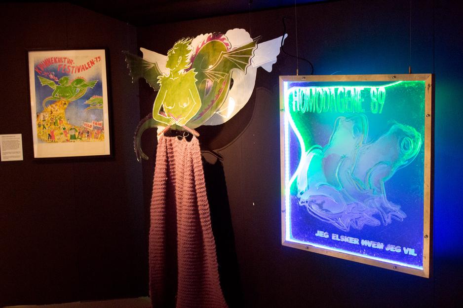 Installasjonen viser en gr&oslash;nn kvinnelig drage Sfinx bl&aring;ser ut ild med teksten "Kvinnekulturfestivalen-79". Hun strikker et gult skjerf  og kvinner i ulike kulturelle aktiviteter er tegnet p&aring; dette skjerfet.