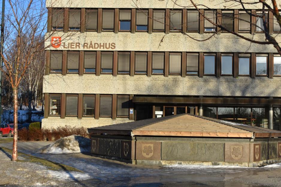 En brun murbygning med ordene "Lier r&aring;dhus" og en r&oslash;dt kommunev&aring;pen med hvite epleblomster i. I front er det en fontene som er lukket for vinteren, samt et lite tre til venstre for denne. 