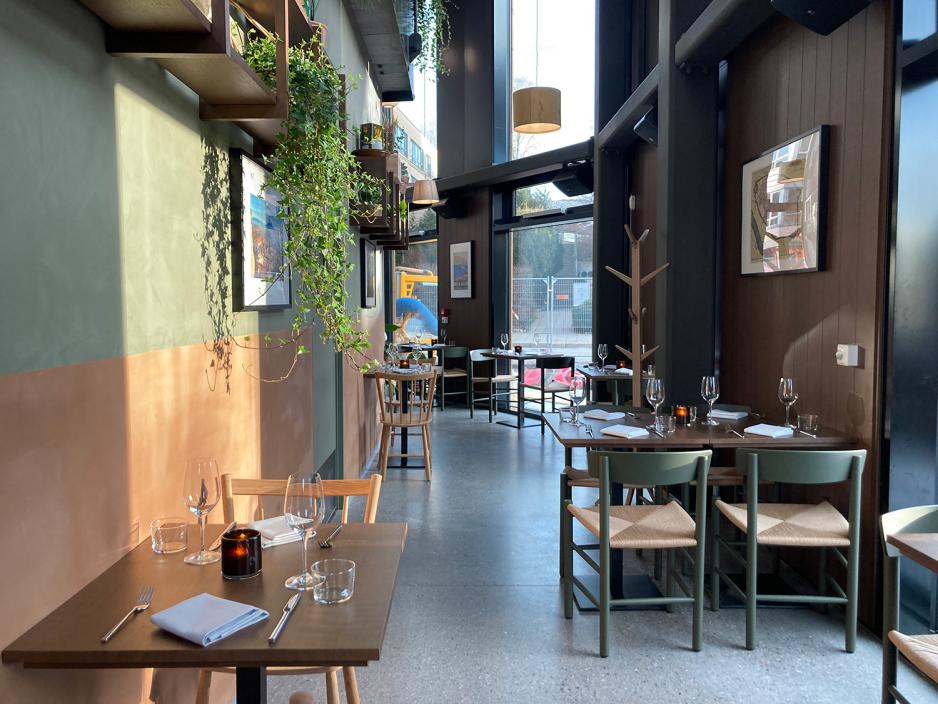 Bildet viser en del av restaurantens lokale. Det er planter, olivengr&oslash;nne og rustenoransje vegger i tillegg til mye lys og sitteplasser i tre. P&aring; bordene er det lys. 