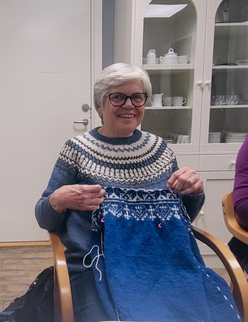 Dame p&aring; strikkekaf&egrave; viser frem den bl&aring; genseren med m&oslash;nster som hun holder p&aring; &aring; strikke.