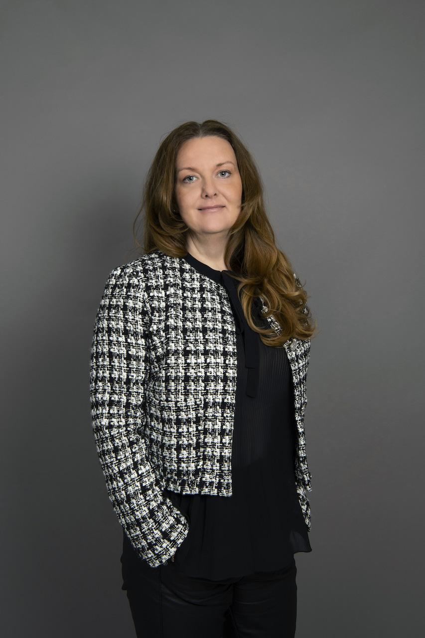 Portrett av Malin Rygg. Hun st&aring;r foran en gr&aring; vegg, og ser inn i kamera med et lite smil. Hun p&aring; svarte kl&aelig;r, og en rutete vevd blazer.