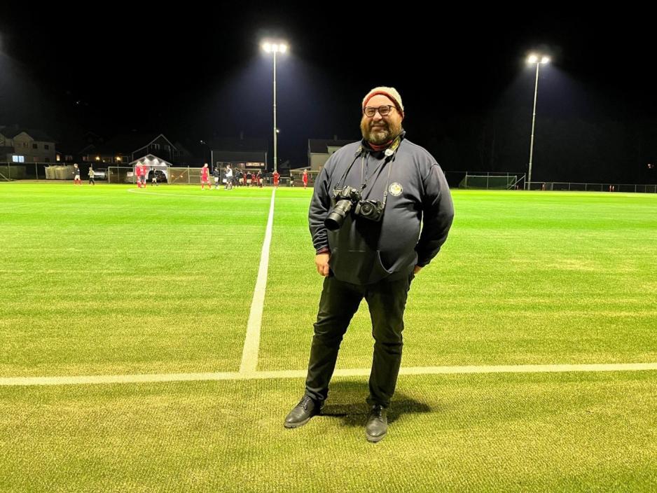 Helbilde av Marius Helg&aring;. Han st&aring;r foran kunstgresset og smiler, med kamera hengende over halsen.