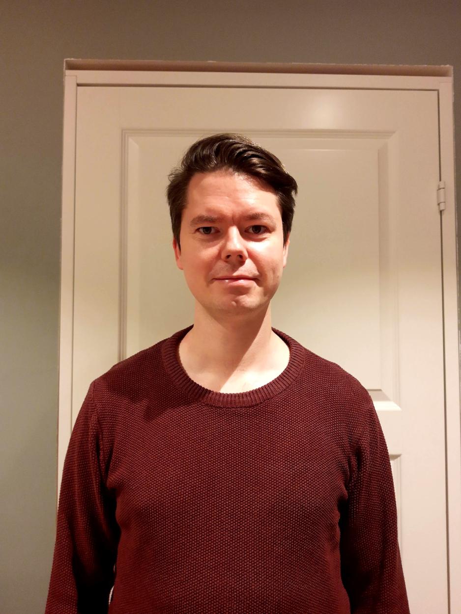 Portrett av mann i tretti&aring;rene som st&aring;r foran en d&oslash;r i burgunderfarget genser.