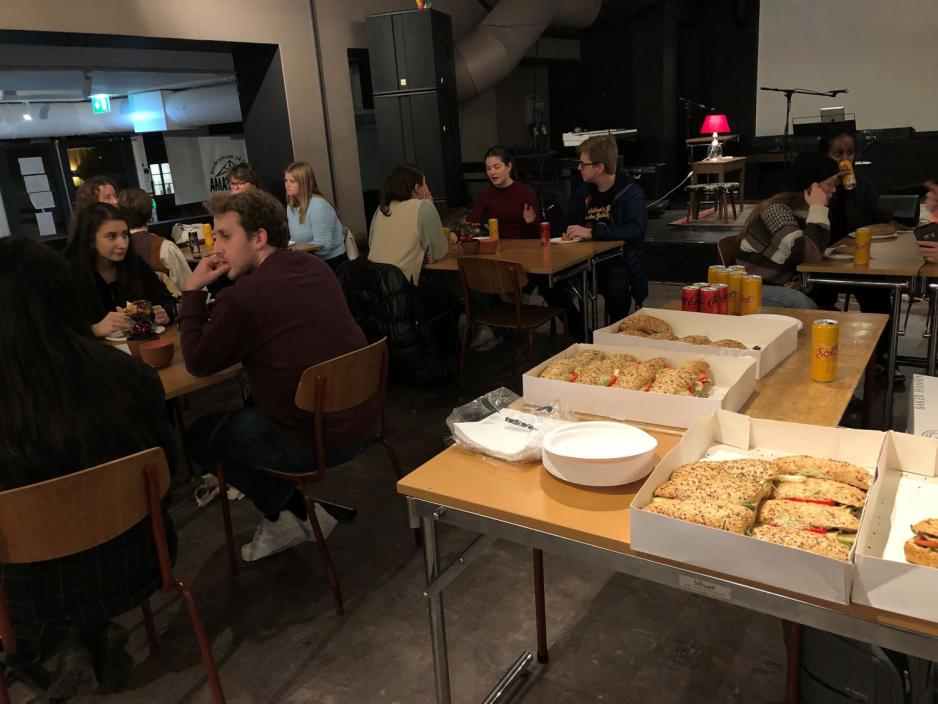 Bord med p&aring;smurte rundstykker. I bakgrunnen sitte mennesker rundt bord og spiser