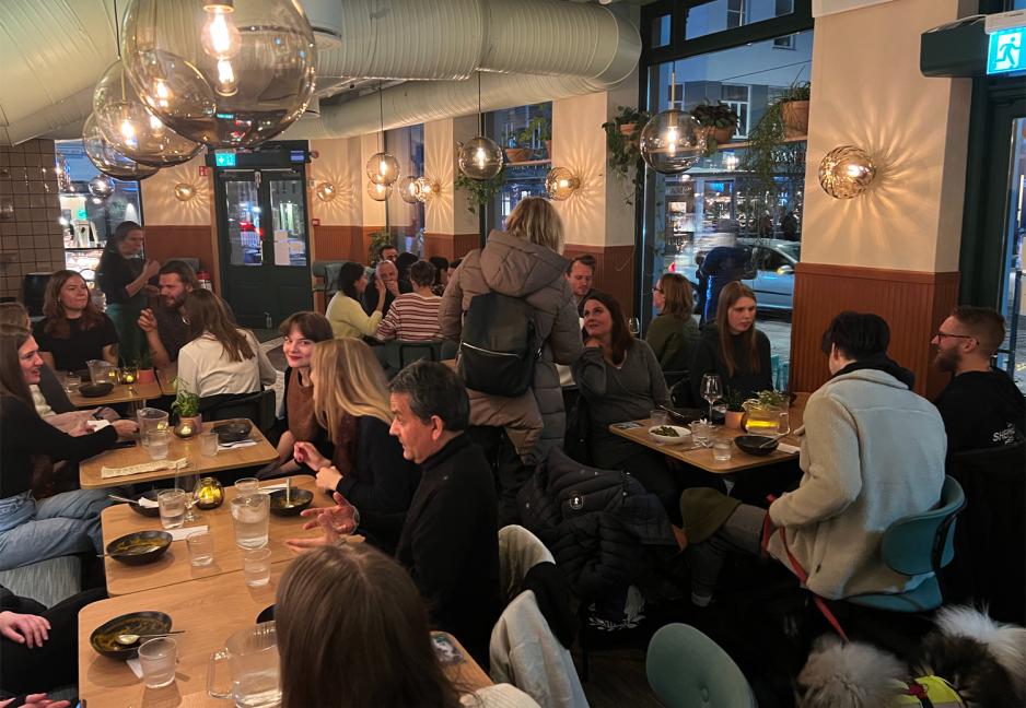 flere titalls personer rundt omkring som sitter og st&aring;r ved bord og spiser mat