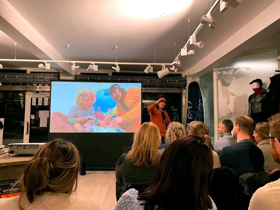 folk h&oslash;rer p&aring; Alexander mens han forteller om turene, p&aring; skjermen ved siden av han er det bilde av mina