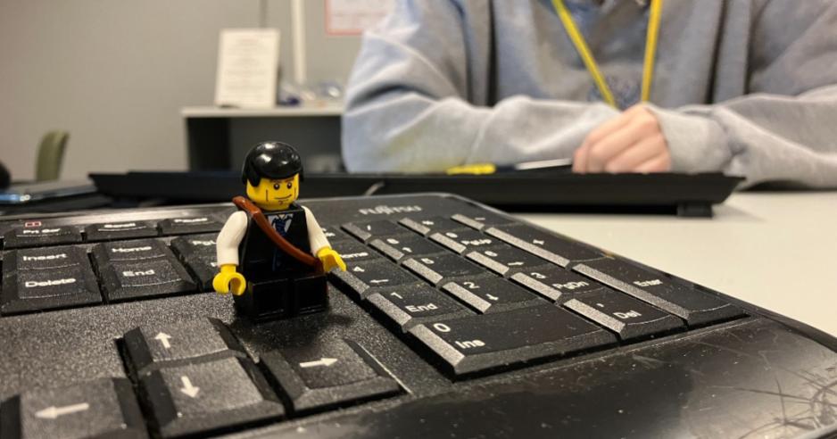 Legomann sittende p&aring; et tastatur (opp&aring; et bord), med overkroppen til en dame og et annet tastatur synlig i bakgrunnen.