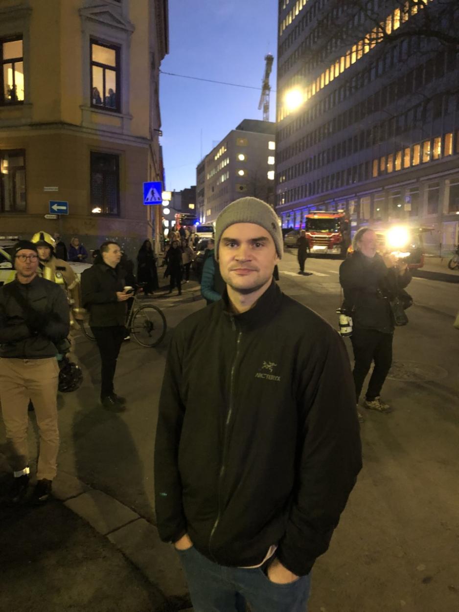 En mann poserer p&aring; fortauskanten, if&oslash;rt gr&aring; lue og fleecejakke, i bakgrunnen st&aring;r en del andre folk og ser p&aring; noe bortenfor til h&oslash;yre/bak kameraet. Det er skumring, en av dem er journalist og tar bilde s&aring; blitzen flasher opp.