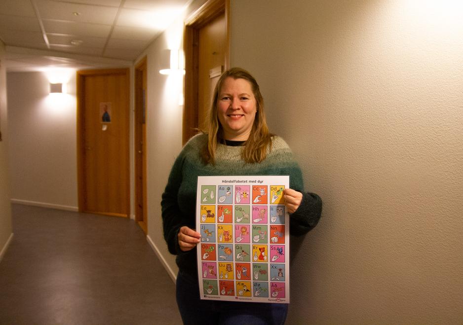 En blond kvinne i gr&oslash;nn genser smiler mot kamera, med en plakat foran seg med tegnspr&aring;kalfabetet. 