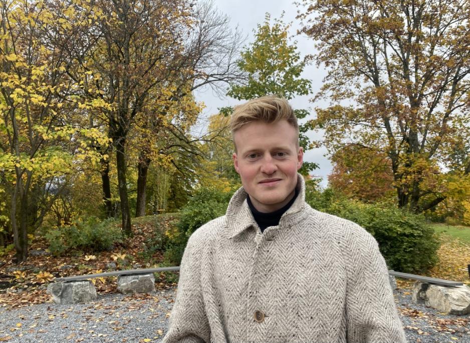 Bilde av en mann i gl&aring; ullk&aring;pe, han har blodt h&aring;r og er ung. Bildet er tatt ute, med tr&aelig;r i bakgrunnen.