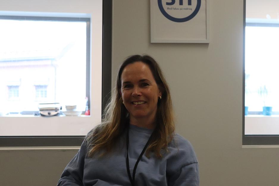 En kvinne sitter foran en vegg og et vindu. Hun har halvlangt h&aring;r og bl&aring; genser.