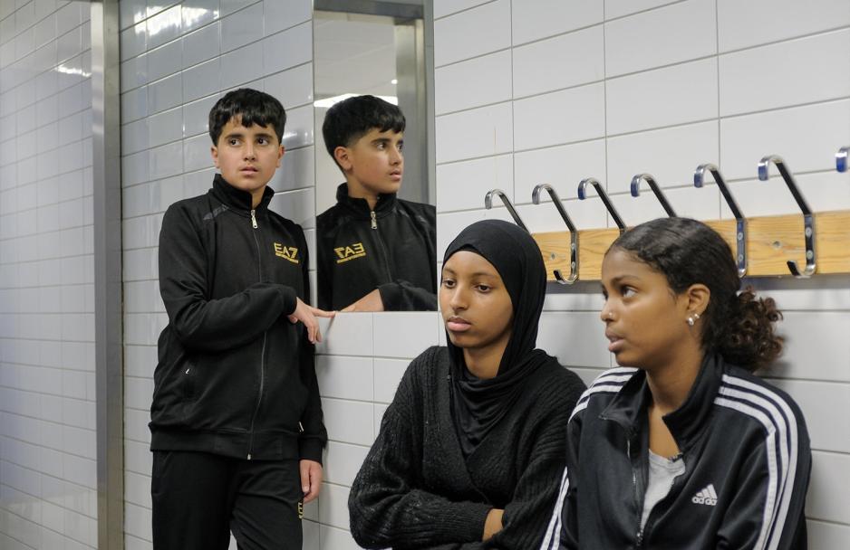 En gutt ser vi to ganger til venstre. B&aring;de i profil og speilbildet hans. De er i garderobe, inntil veggen . Vi ser to jenter som sitter nede til h&oslash;yre for gutten.