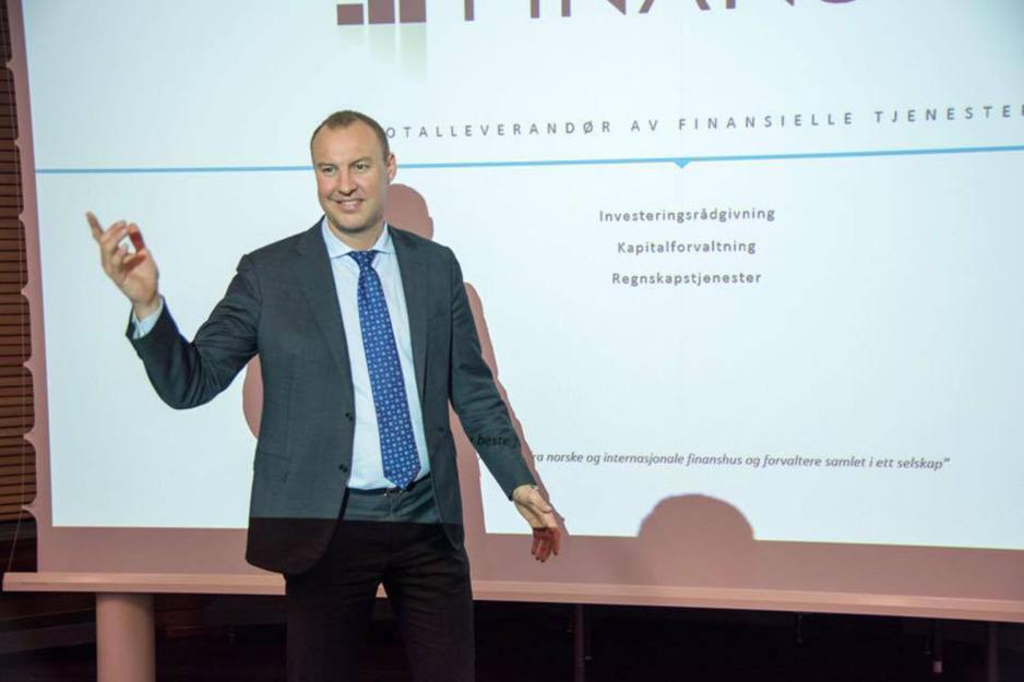 En mann i dress st&aring;r foran en skjerm og gestikulerer.