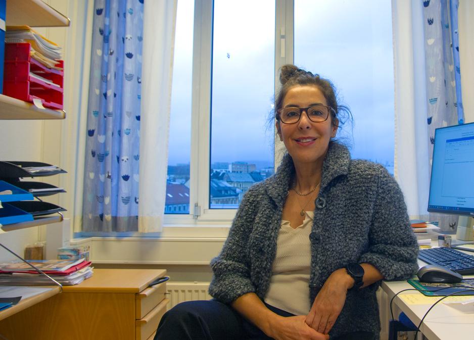 En dame med m&oslash;rkt h&aring;r samlet i en dott, smiler til kamera med store vinduer bak seg med utsikt utover et t&aring;kete Oslo. 