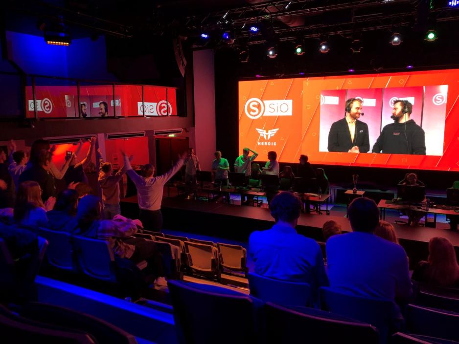 Mange personer st&aring;r oppreist og heier p&aring; en tribune. Foran dem st&aring;r spillerne