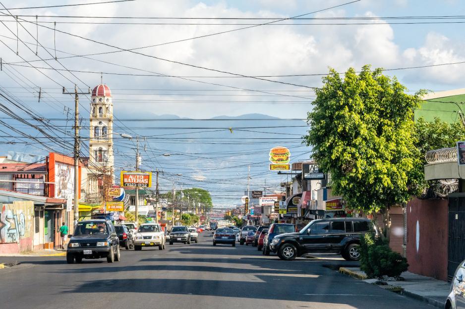 Trafikkert vei i San Jos&eacute; i Costa Rica. Mange biler, lave forretningslokaler og tet-i-tett med telefonledninger.