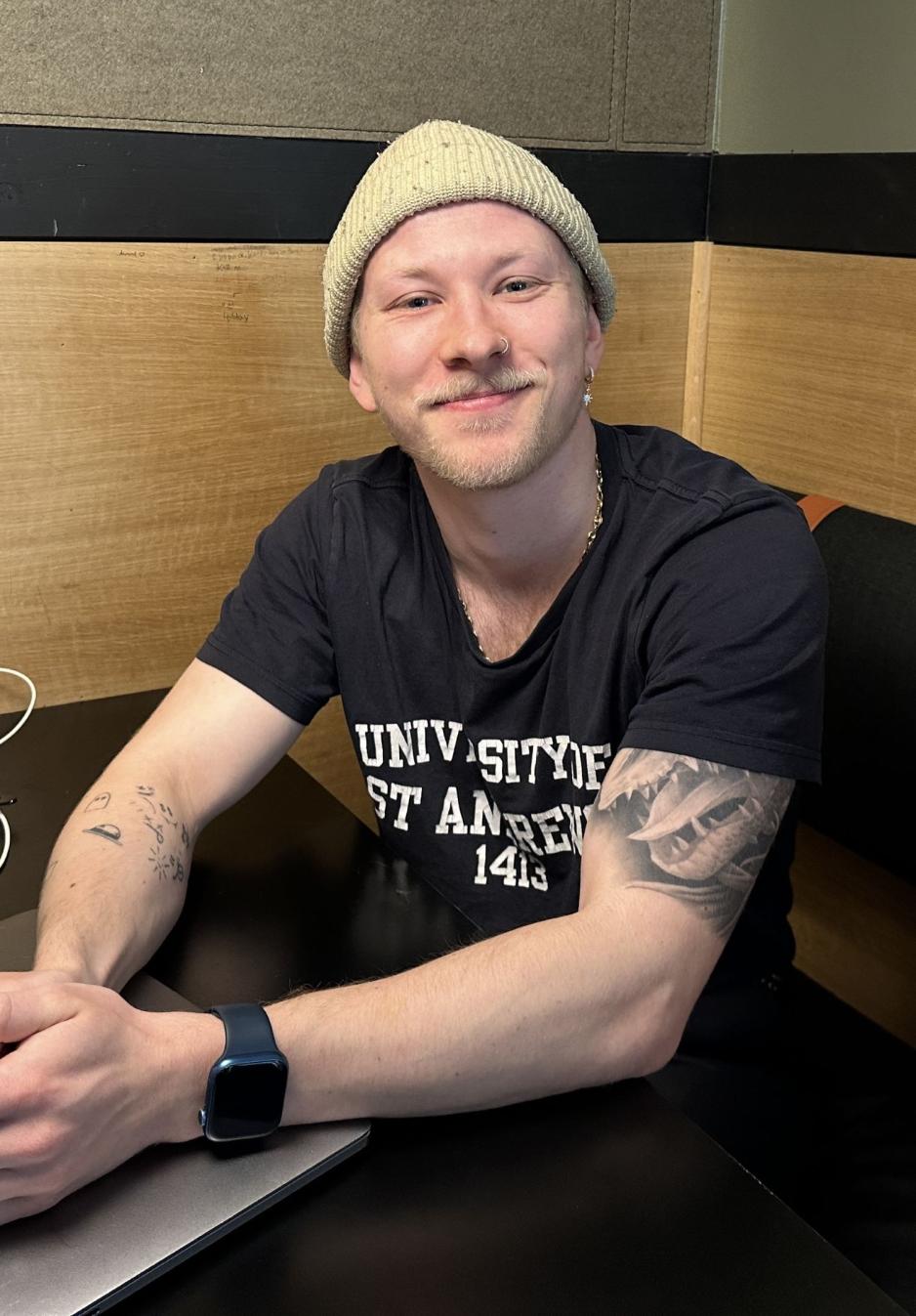 En smilende gutt med lue og piercing i nesa smiler til kamera, mens han hviler albuene p&aring; bordet. 