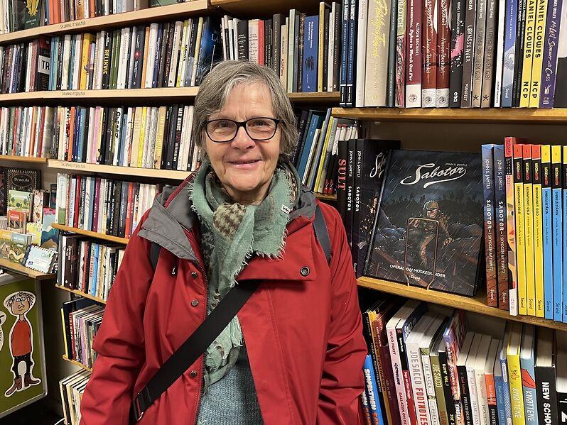 Sissel S&oslash;rensen har gr&aring;tt h&aring;r og briller og st&aring;r smilende foran en bokhylle inne p&aring; Sagene Bokhandel. 