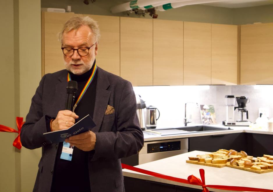 Odd Rune Larsen st&aring;r med mikrofon foran et r&oslash;dt b&aring;nd ved kj&oslash;kkenet i lokalet og holder tale. 