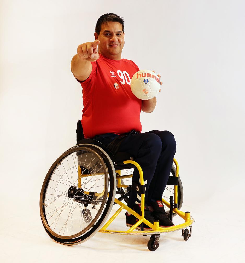 Tor Solstad sitter i en rullestol og peker mot kamera med en ball i den andre h&aring;nden. Han er i et fotostudio