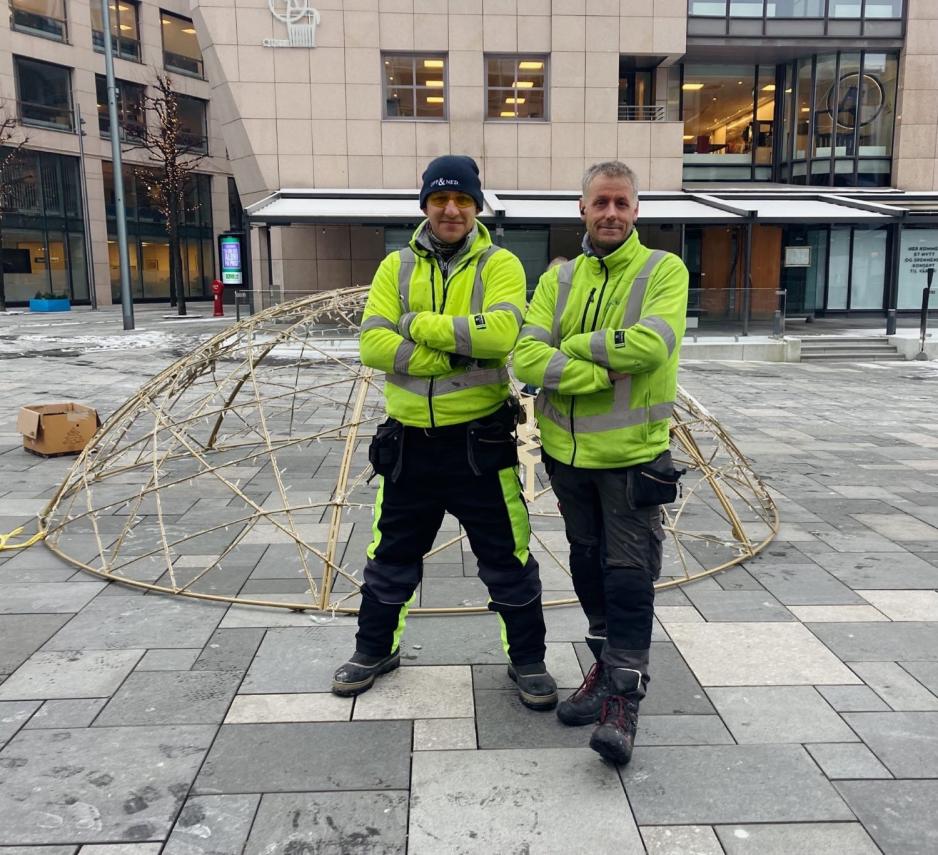 To menn i gr&oslash;nne jakker st&aring;r foran den demonterte julekulen p&aring; Bryggetorget.