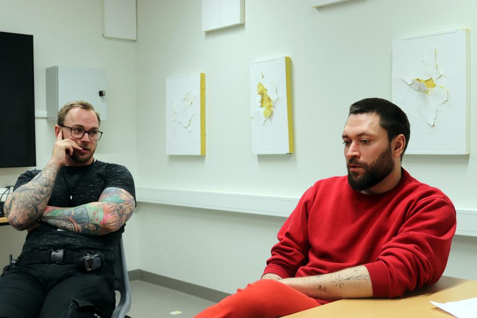 En mann i r&oslash;d genser og joggebukse sitter ved et bord. Ved siden av han sitter en fengselsbetjent i t-skjorte og tatoverte armer.