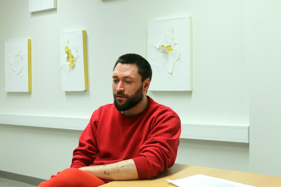 En mann i tretti&aring;rene med m&oslash;rkt h&aring;r og skjegg sitter ved et bord i en r&oslash;d genser og r&oslash;d bukse.