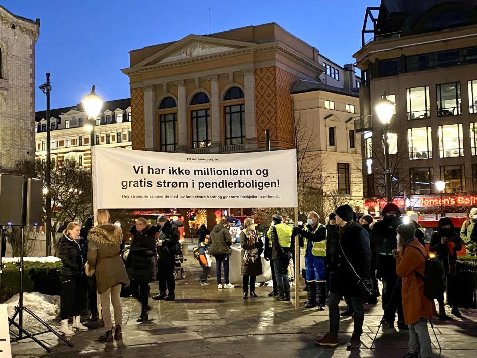 To menn holder et stort banner med teksten "Vi har ikke millionl&oslash;nn og gratis str&oslash;m i pendlerboligen". Flere demonstranter vrimler rundt dem. 