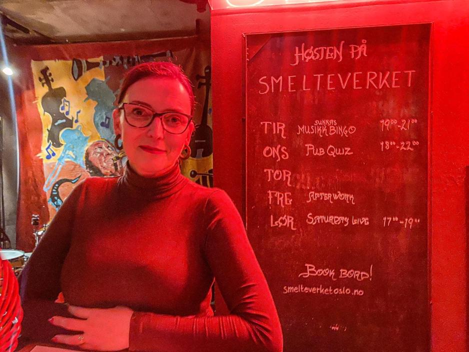 Her er Sunna, ei dame som har p&aring; seg briller, h&aring;ret i hestehale og smiler til kameraet. Hun har p&aring; seg en lang hals genser og st&aring;r med hendene samlet. 