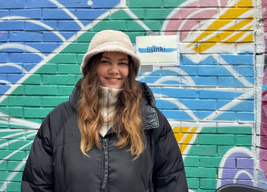 Bildet viser en smilende Sunniva Kiste Bergan som stiller hver s&oslash;ndag med eget h&aring;ndarbeid p&aring; markedet i Ingensgate. Det er fargerik bakgrunn preget av graffiti.