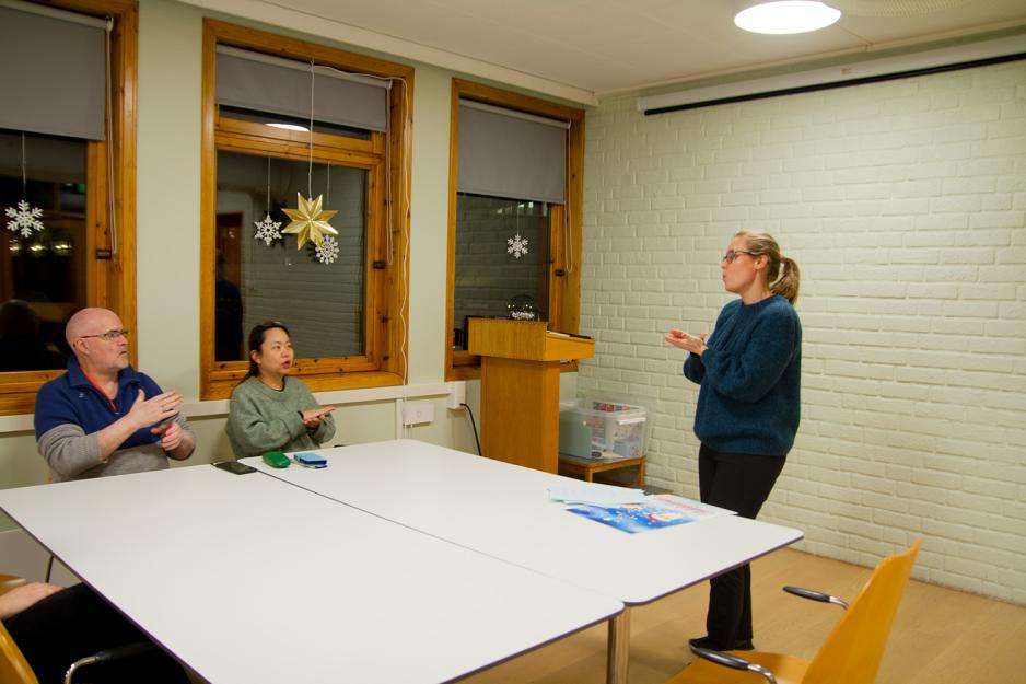 Bildet viser tre personer, to som sitter ved et bord, og ei st&aring;r foran bordet. Alle viser tegnspr&aring;ktegnet for kakao. 