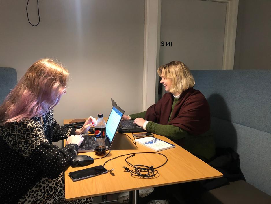 To jenter sitter ved et bord og skriver p&aring; hver sin datamaskin.