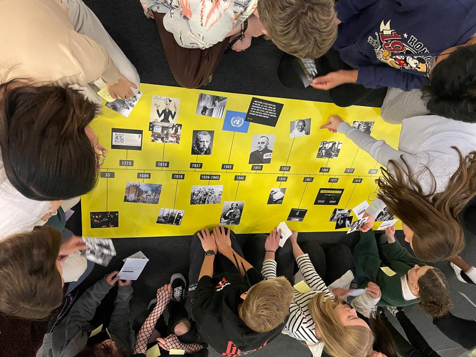 Ungdommer sine hender som plasserer lappet med historiske begivenheter p&aring; p&aring; en stor gul tidslinje.