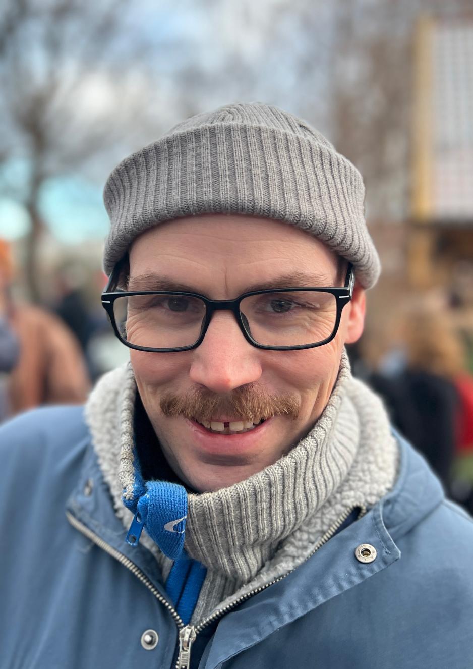 Mann med briller og gr&aring; lue, med stor brun og fyldig bart