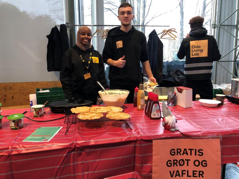 En jente og en gutt st&aring;r smilende bak et bord. P&aring; bordet er et vaffeljern og en kjele med gr&oslash;t