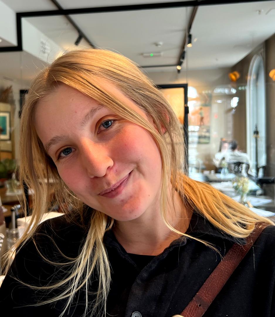 Portrettbilde p&aring; en cafe av V&aring;rin Strand med blondt h&aring;r og svart frakk, skinnstropp synelig langs skulderen med hode p&aring; skr&aring;tt og et smil