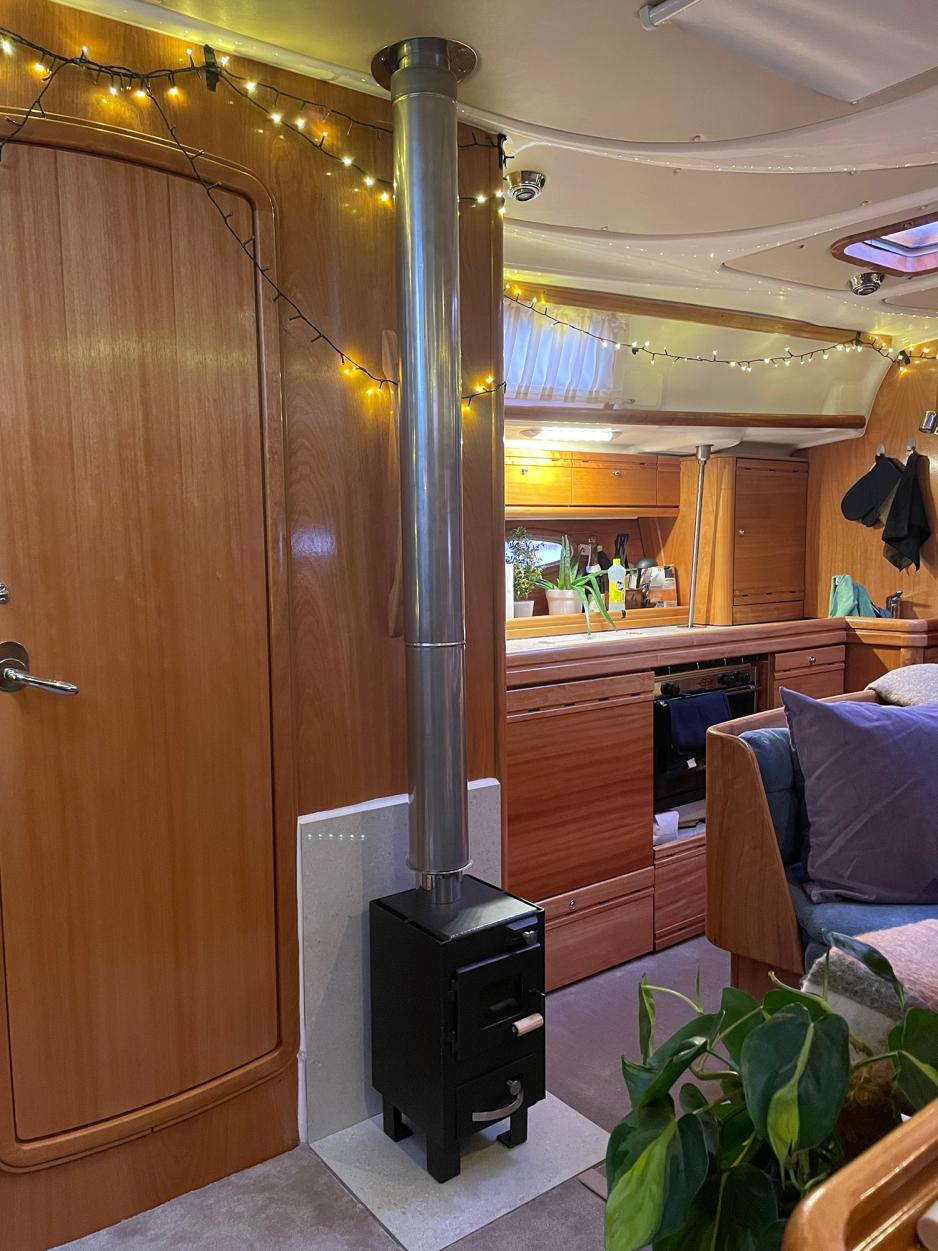 Liten svar vedovn med et langt r&oslash;r i rustfritt st&aring;l som g&aring;r opp gjennom taket. Vedovnen er plassert p&aring; en gr&aring; flis i seilb&aring;ten. Den brune treveggen bak ovnen er beskyttet med den samme gr&aring;e flisen som er brukt p&aring; gulvet. 