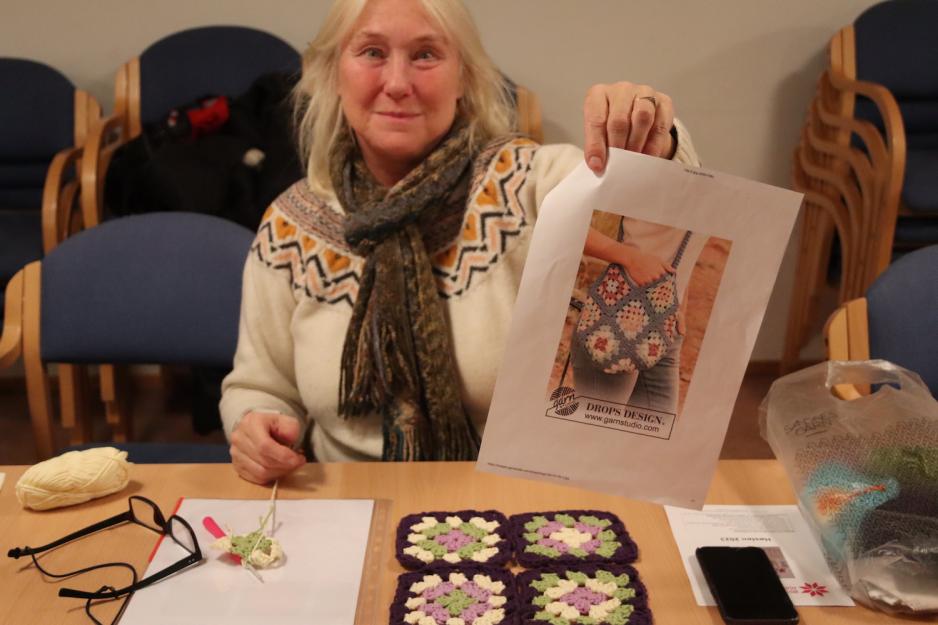 Dame viser frem bilde av en veske og har ulike h&aring;ndarbeid foran seg p&aring; bordet