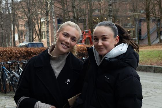 To damer som st&aring;r vedsiden av hverandre og smiler, med en plen i bakgrunnen