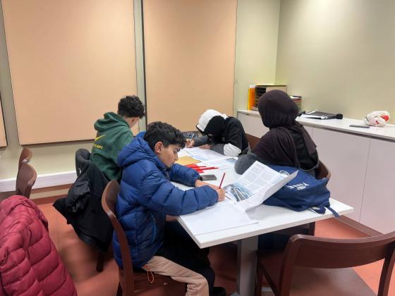 Tre gutter sitter ved et bord og gj&oslash;r lekser sammen med lederen av leksehjelp. 