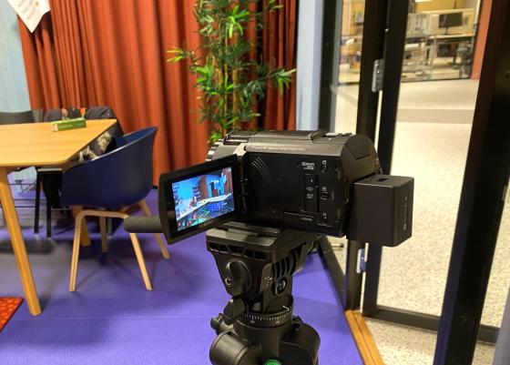 Et filmkamera som filmer to personer, som sitter og snakker p&aring; et fargerikt m&oslash;terom.