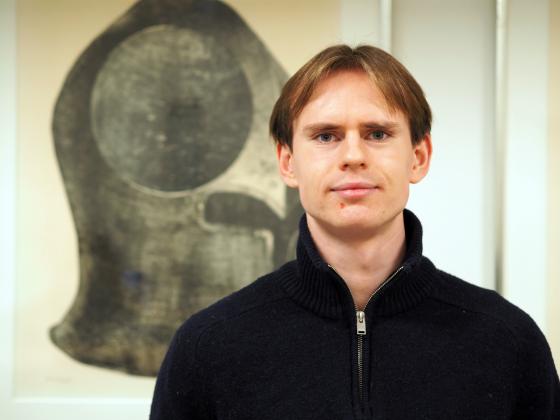 Mann st&aring;r foran et svart maleri, han har p&aring; seg en m&oslash;rkebl&aring; genser.