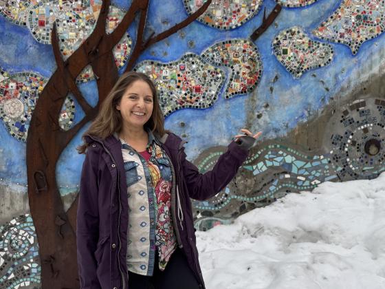 Jannicke Wiel st&aring;r foran en vegg dekorert med fargerike steiner i m&oslash;nster av et tre. Smiler og l&oslash;fter armen for &aring; vise frem. 