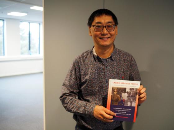 Man st&aring;r foran en gr&aring; vegg. Han smiler og viser fram en bok.