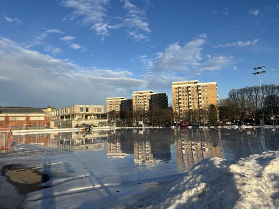 En stor isflate med masse vann opp&aring;. P&aring; isen kan man se en traktor som kj&oslash;rer. I bakgrunnen er det h&oslash;ye leilighetsblokker. Solen skinner over vannoverflaten p&aring; isen.