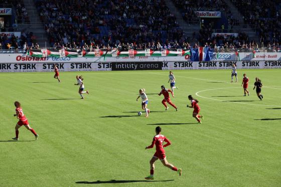To fotballlag spiller mot hverandre p&aring; en kunstgressbane. Det palestinske laget har r&oslash;d drakter. V&aring;lerenga sitt lag har hvite drakter. Det er tilskuere p&aring; tribunen.  