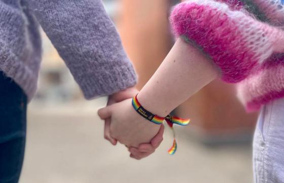 Bildet viser to personer som holder hender. Ene parten har p&aring; seg et regnbuearmb&aring;nd.