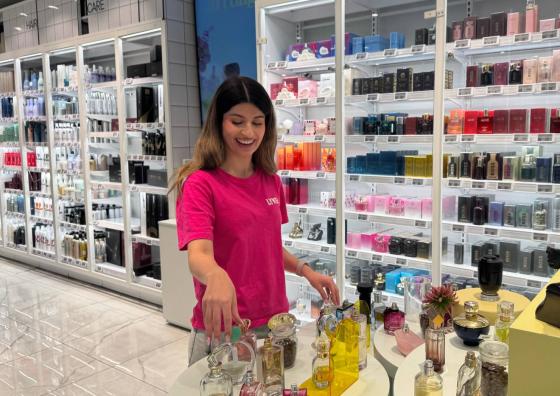 jente med stort smil m&oslash;rkt h&aring;r og rosa t skjorte fikser p&aring; parfymeflasker p&aring; et hvitt bord